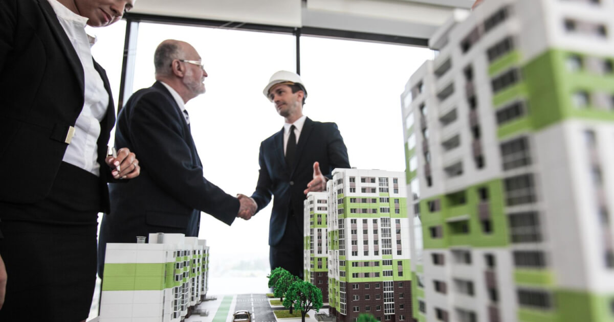 Two professionals shaking hands over a detailed pre-construction model, symbolizing a successful sale in sustainable urban development.