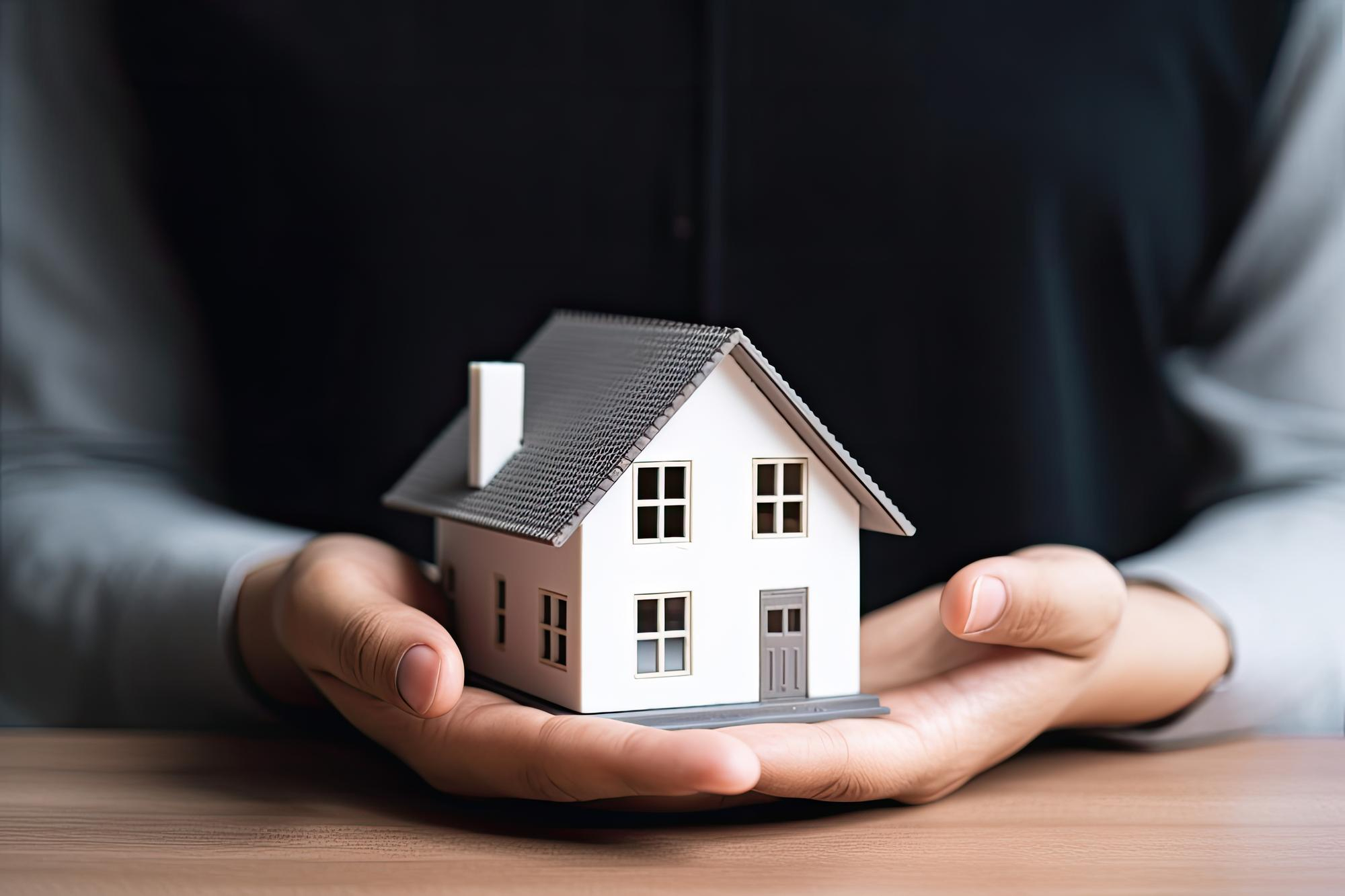 A real estate agent, dressed in a black shirt, holding a small model of a white house with a gray roof in their hands.
